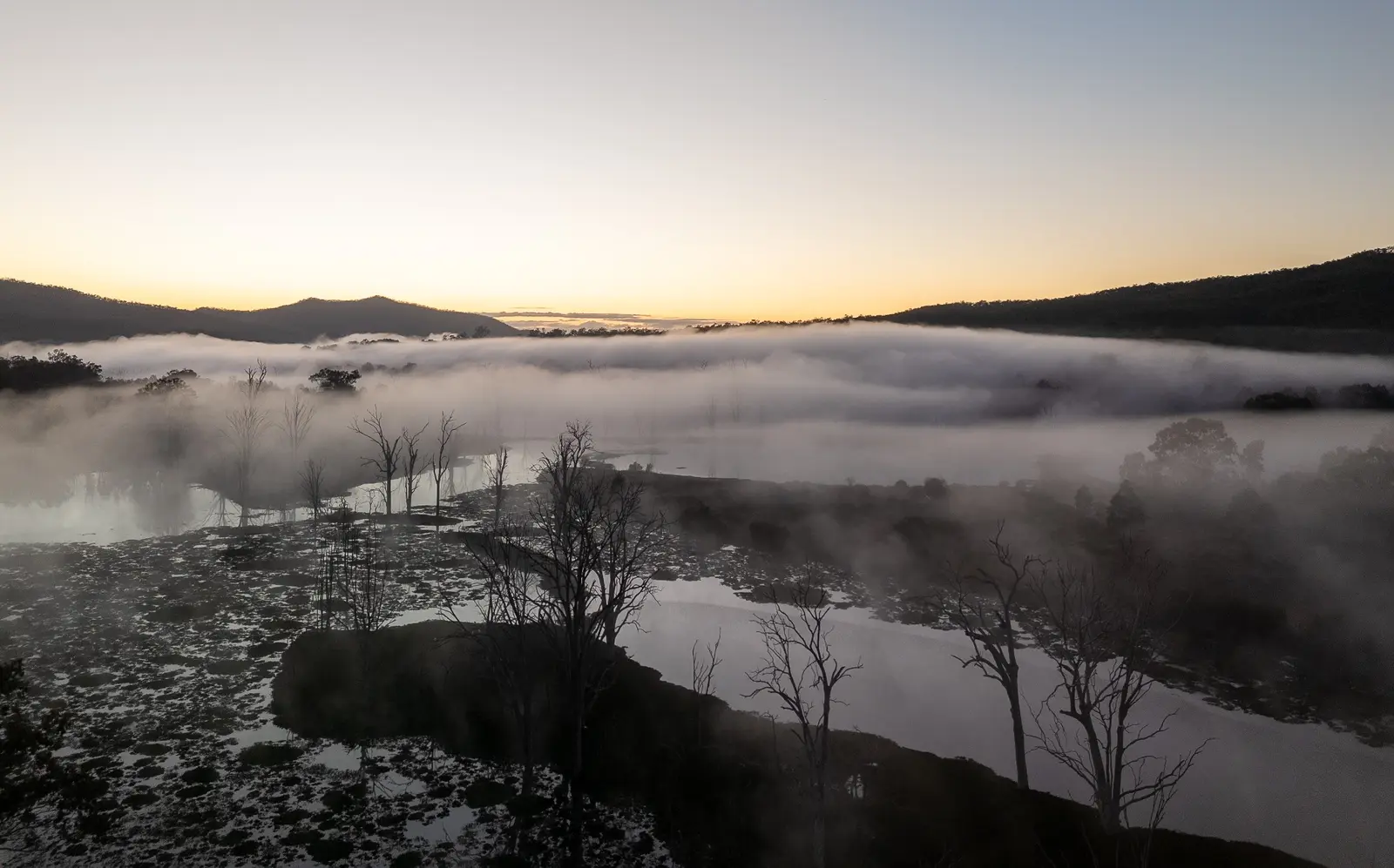 Mist drifting across a lake at sunrise, with bare trees reflected in the water